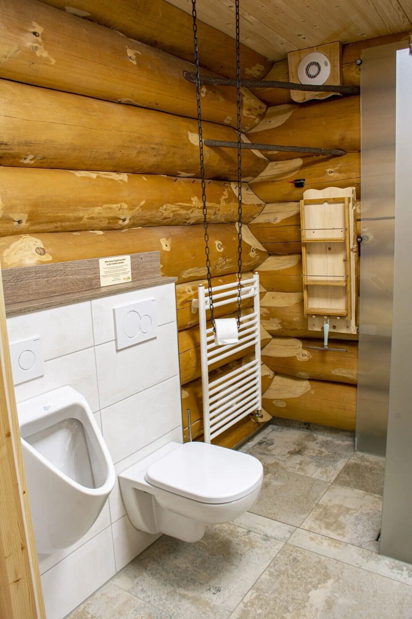 A wooden cabin bathroom with a toilet, urinal, and a hanging towel rack.