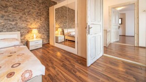 A bedroom with a stone wall, wooden flooring, a bed with a patterned bedspread, bedside lamps, a wardrobe with a mirror, and an open door leading to another room.