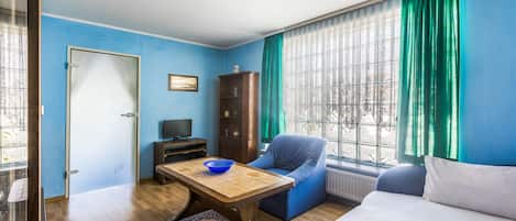 Une pièce avec une table en bois, une chaise bleue, un canapé, un meuble et une fenêtre avec des rideaux.