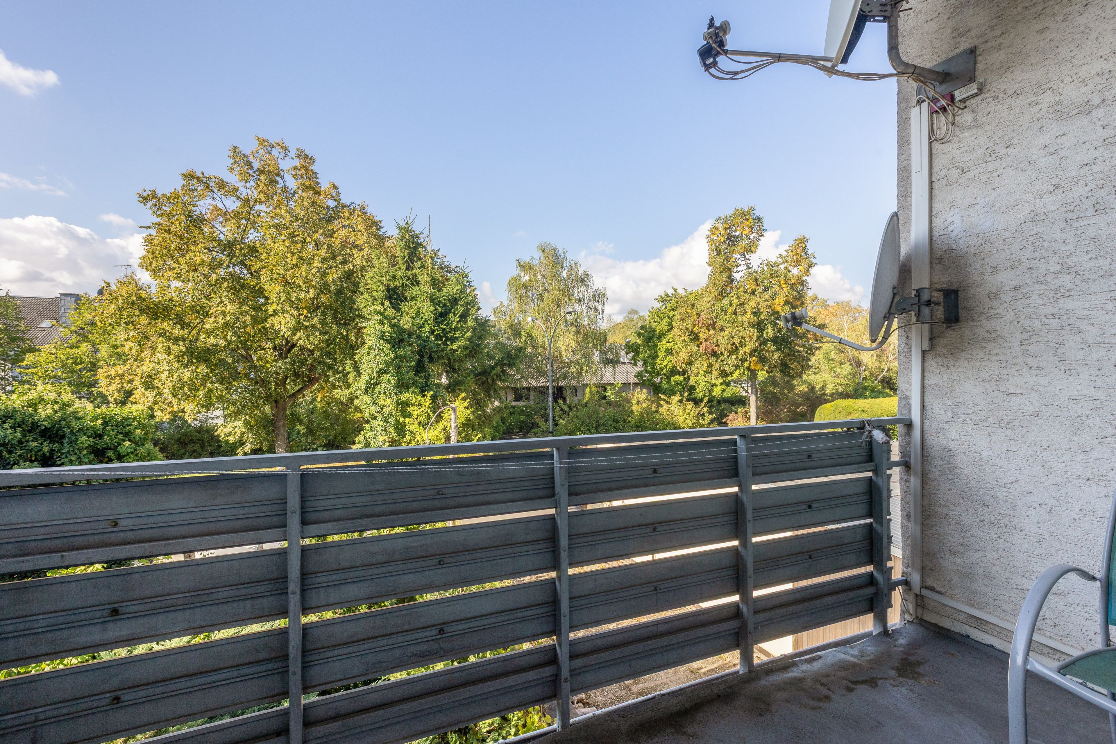 Ein Balkon mit Metallgeländer, einer an der Wand befestigten Satellitenschüssel und Blick auf Bäume und Gebäude im Hintergrund.