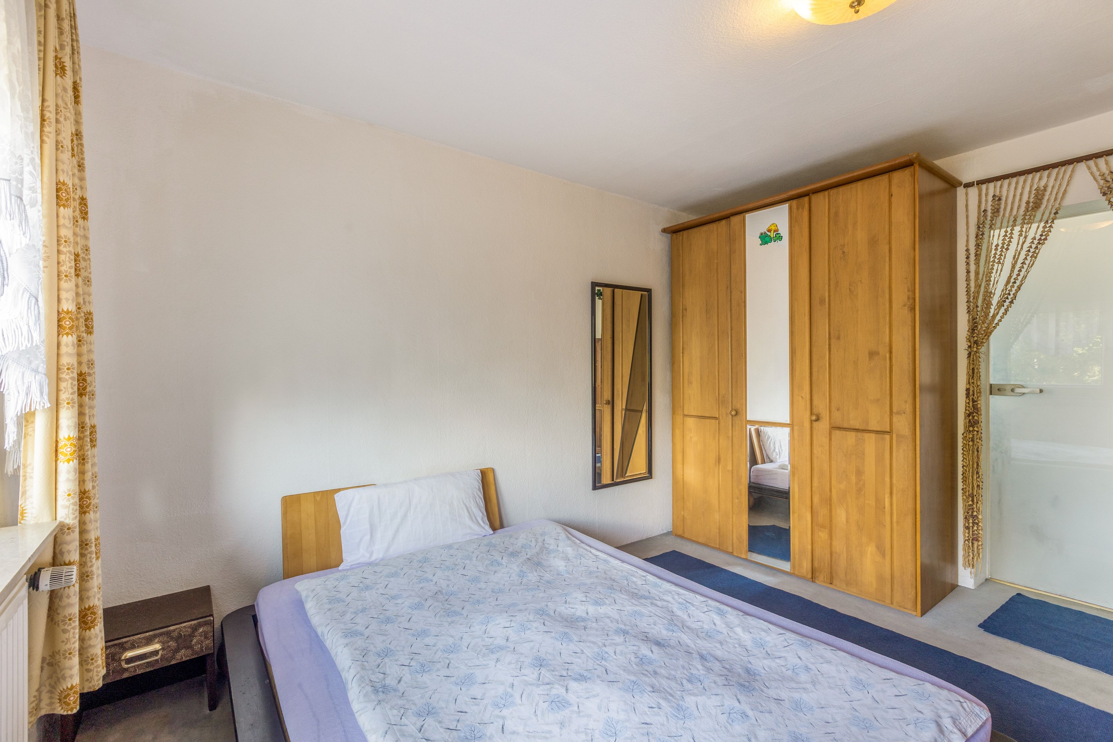 Ein Schlafzimmer mit Bett, Kleiderschrank, Spiegel und einem Fenster mit Vorhängen.