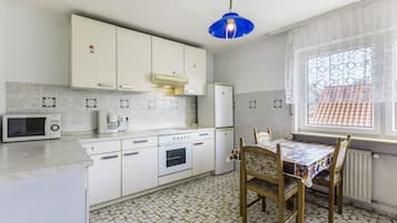 Eine kompakte Küche mit weißen Schränken, einer Mikrowelle, einem Herd und einem Kühlschrank. Es gibt einen Esstisch mit Stühlen und ein Fenster mit gemusterten Vorhängen.