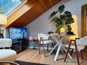 A cozy living room with a TV, a dining table, chairs, a large plant, and a view through a skylight.