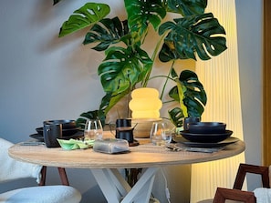 Une table ronde en bois, agrémentée de verres noirs et transparents, d’une lampe jaune et d’une grande plante verte en arrière-plan.