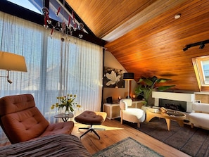 A modern living room with a wooden ceiling, a large window with sheer curtains, a brown leather armchair, a white sofa, a small round table, and a floor lamp.