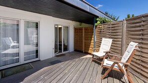 Una terraza de madera con dos sillas blancas acolchadas, un gran ventanal corredero y una valla de madera.