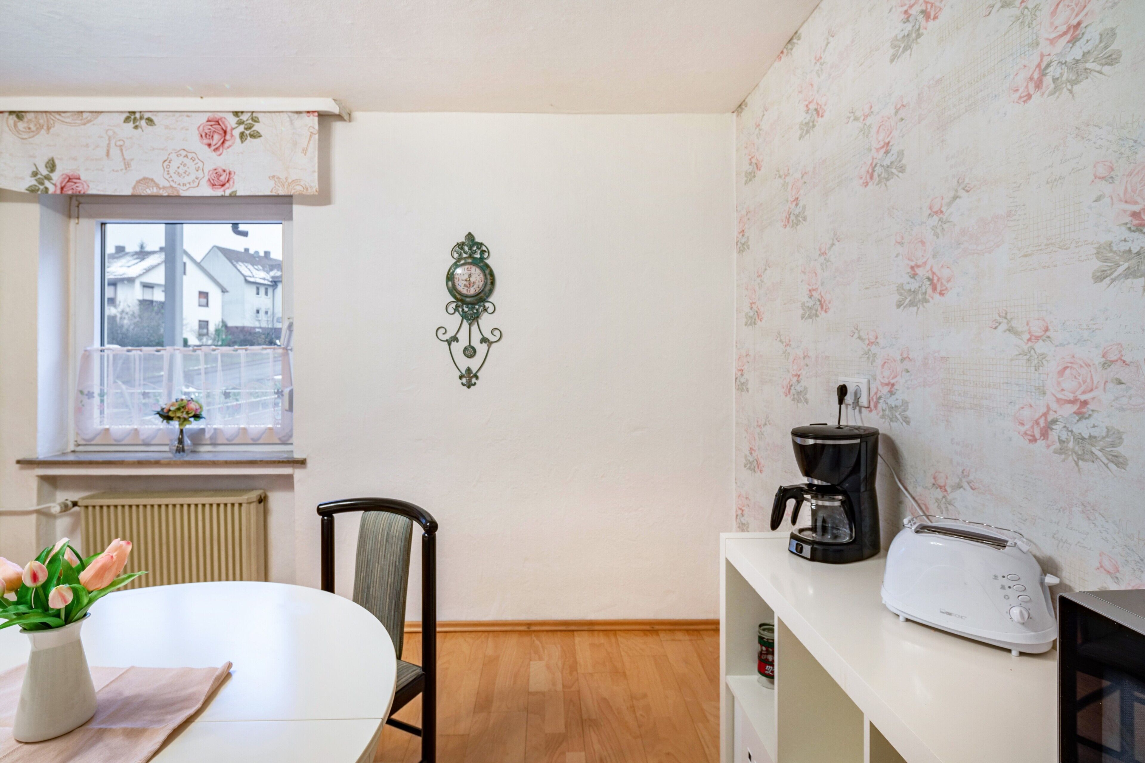 Una cocina con una mesa blanca, una silla, un tostador, una cafetera y un reloj de pared decorativo.