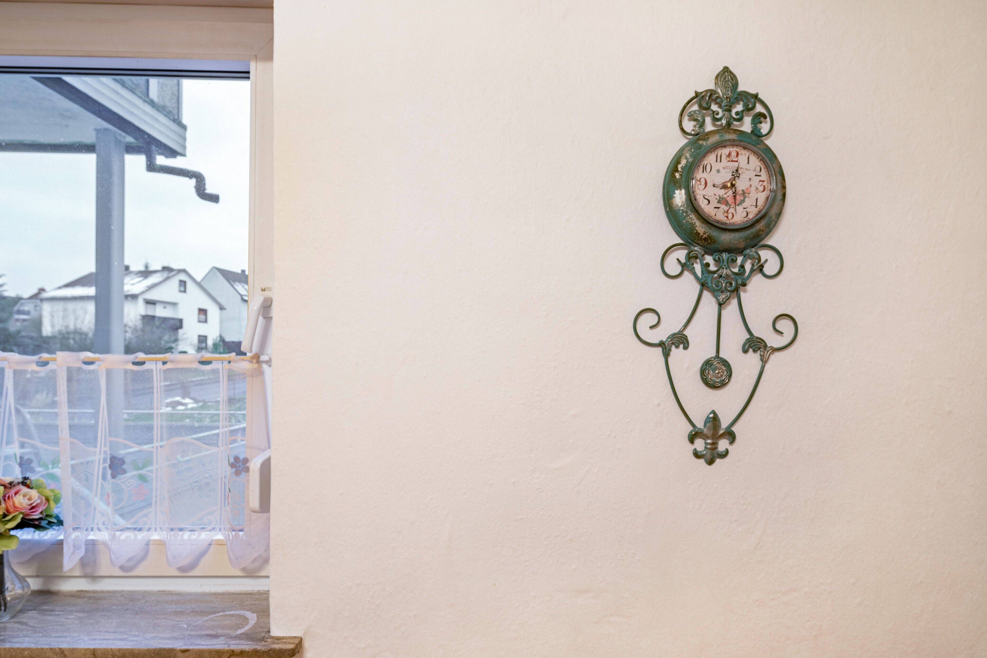 Une horloge murale dotée d’un cadre décoratif, une fenêtre donnant sur des maisons et un rideau orné de motifs floraux.