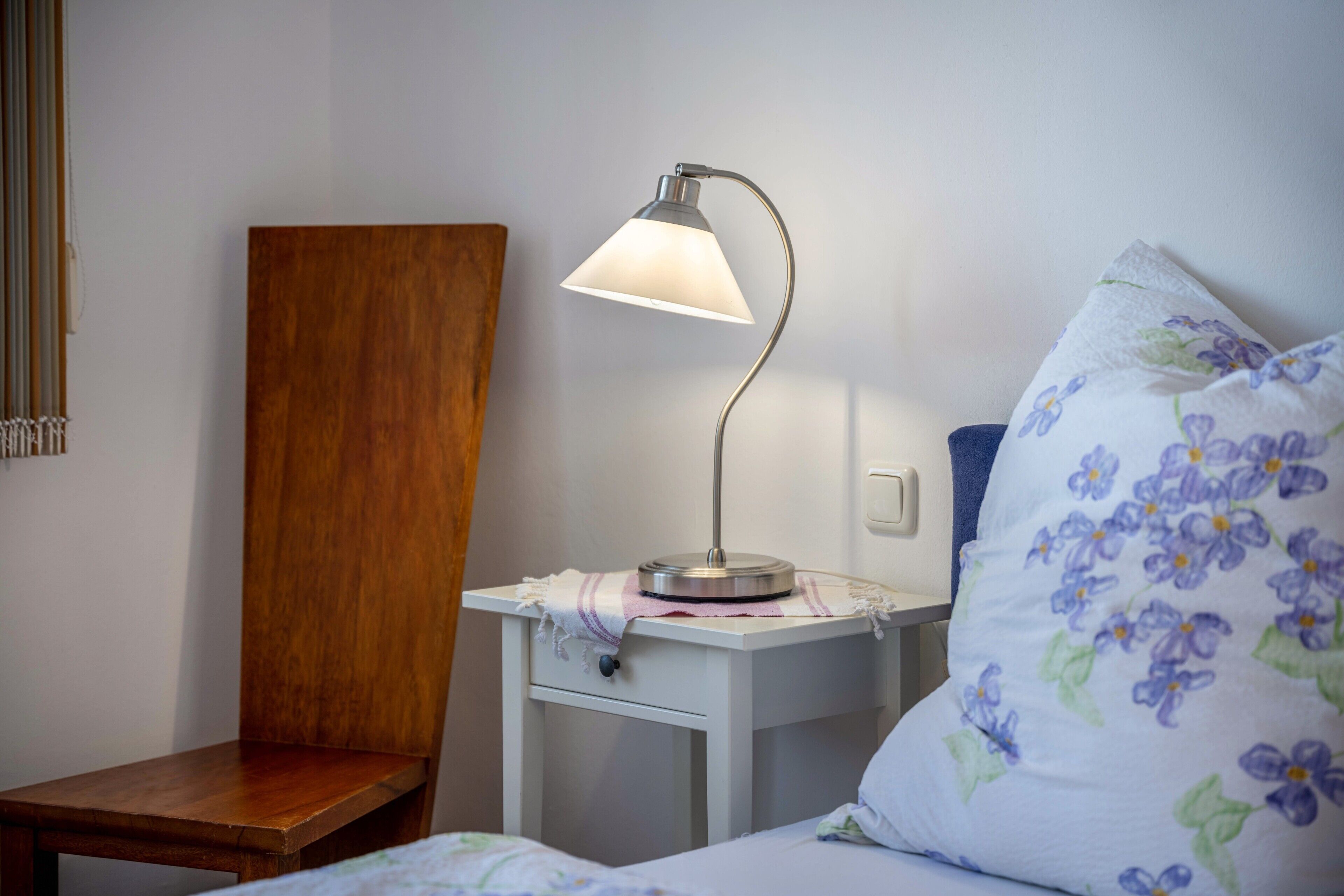 A bedroom with a wooden chair, a bedside table with a lamp, a floral pillow, and a bed with a white sheet.