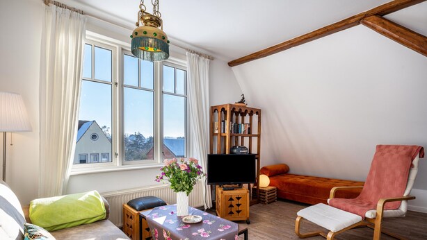 Una sala de estar acogedora con un sofá, una silla, una mesa con un florero, un televisor sobre un mueble de madera y vistas a través de un ventanal.