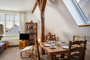 A dining area with a wooden table set for four, a television, a bookshelf, and a window with curtains.