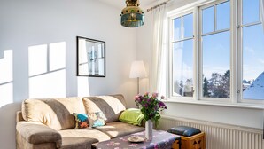 Un salón con sofá, una mesa con mantel floral, un jarrón con flores, una lámpara y una ventana que ofrece vistas a casas.