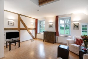 Uma sala de estar com televisão, vigas de madeira, um sofá branco, um armário e janelas amplas com cortinas.
