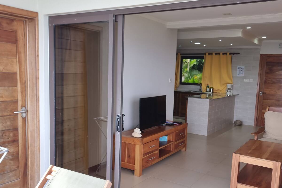 A modern living room with a sliding glass door leading to a balcony, a wooden TV stand, and a small dining area.