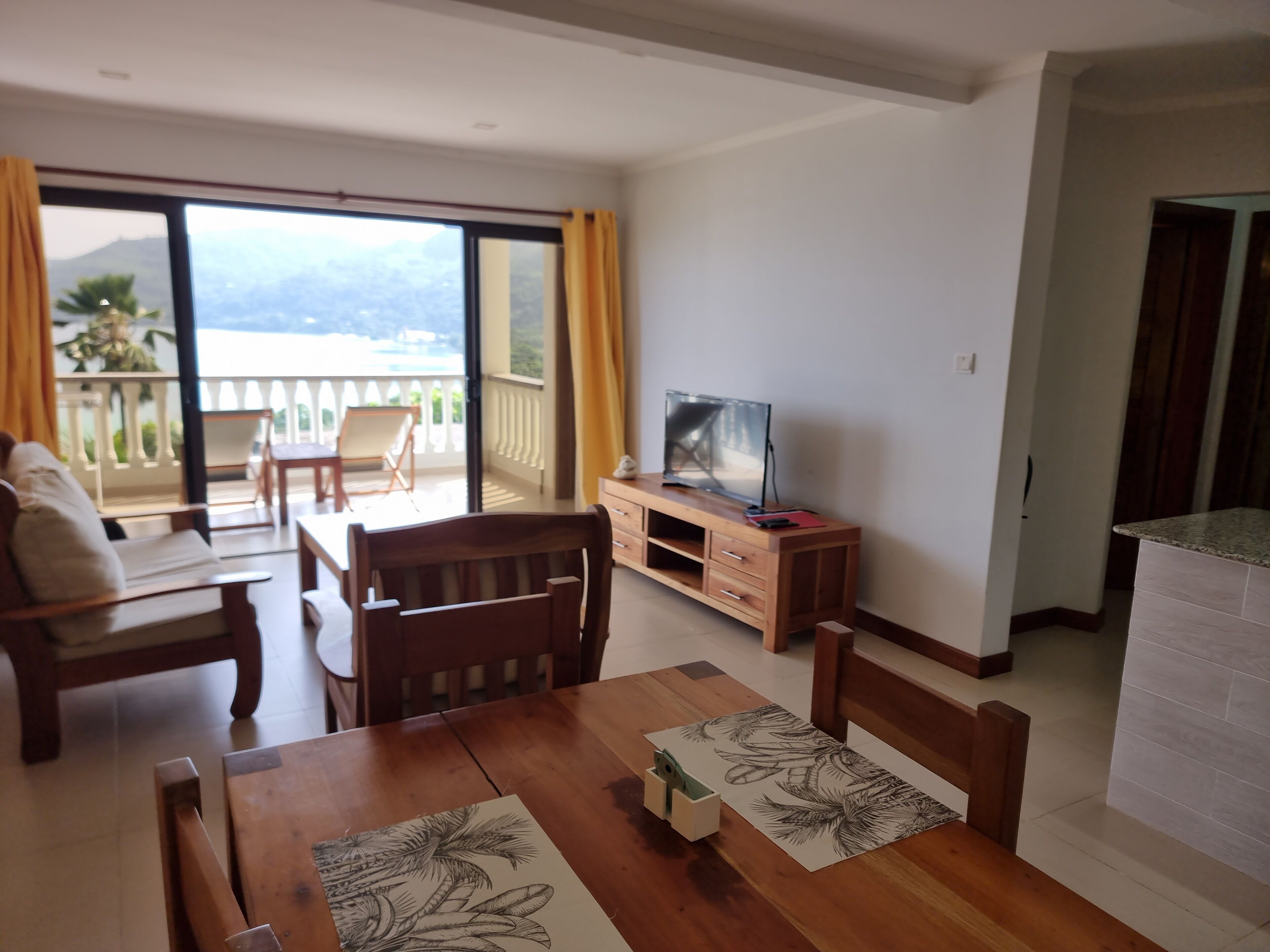 Una habitación con una mesa de comedor de madera, sillas, un televisor sobre un mueble de madera y un balcón con vistas a las montañas.