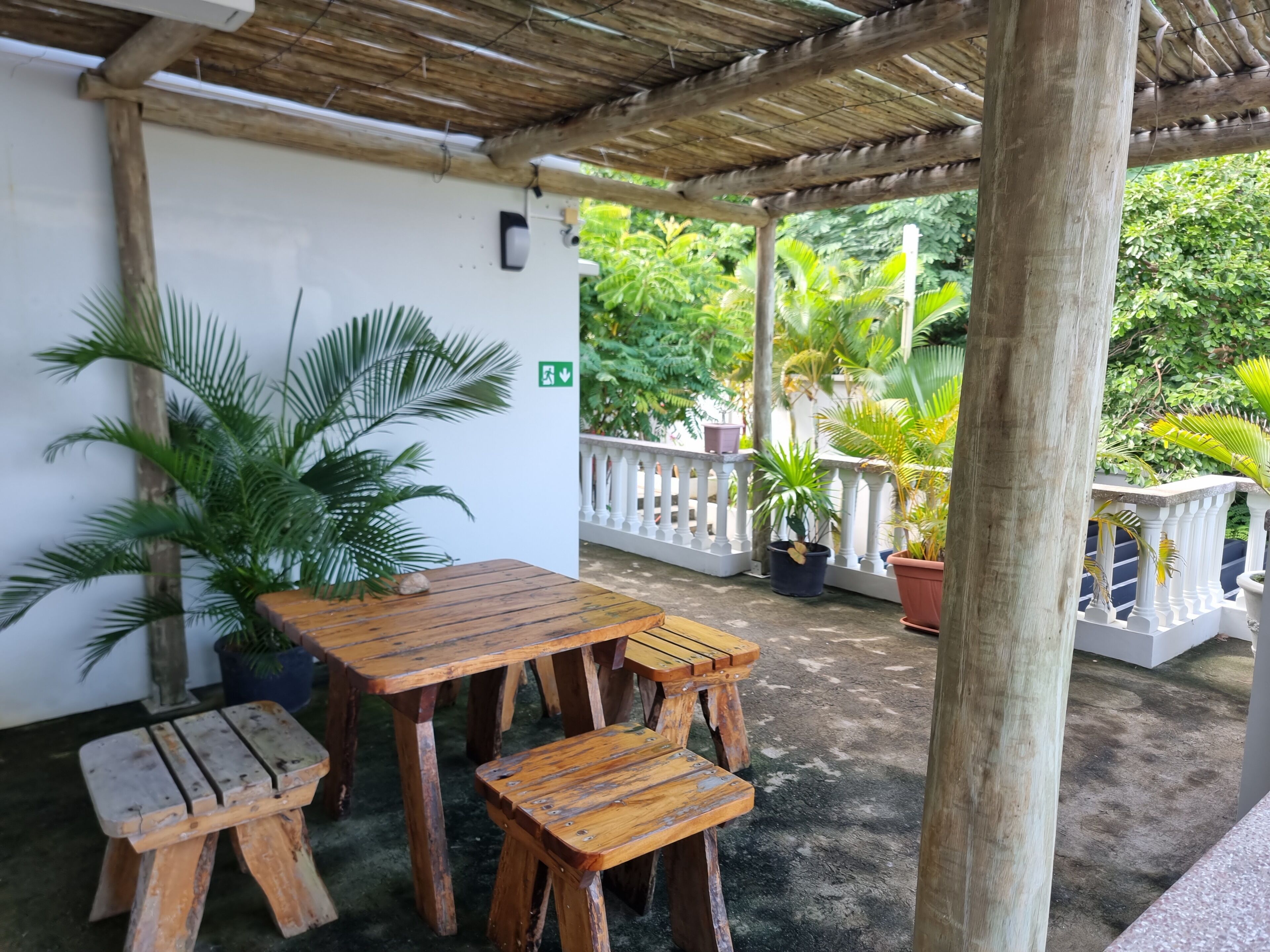 Un espace extérieur aménagé avec des meubles en bois, comprenant une table et des bancs, entouré de plantes en pot et d’une rambarde blanche.