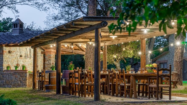 Un espacio exterior para comer con mesas y sillas de madera, paredes de piedra y luces colgantes.