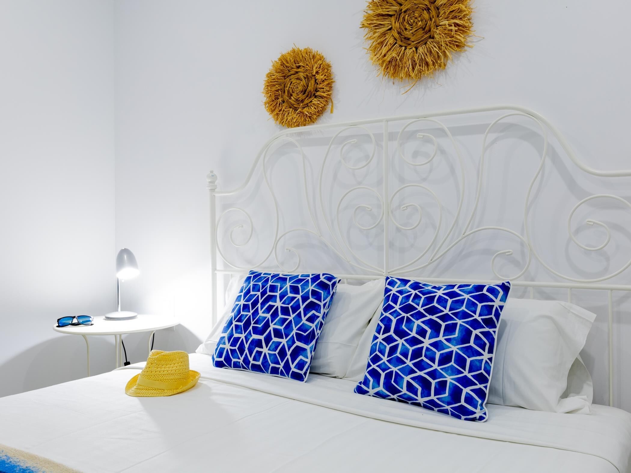 A neatly made bed with white bedding and blue patterned pillows, a small bedside table with a lamp, and a yellow hat on the bed.