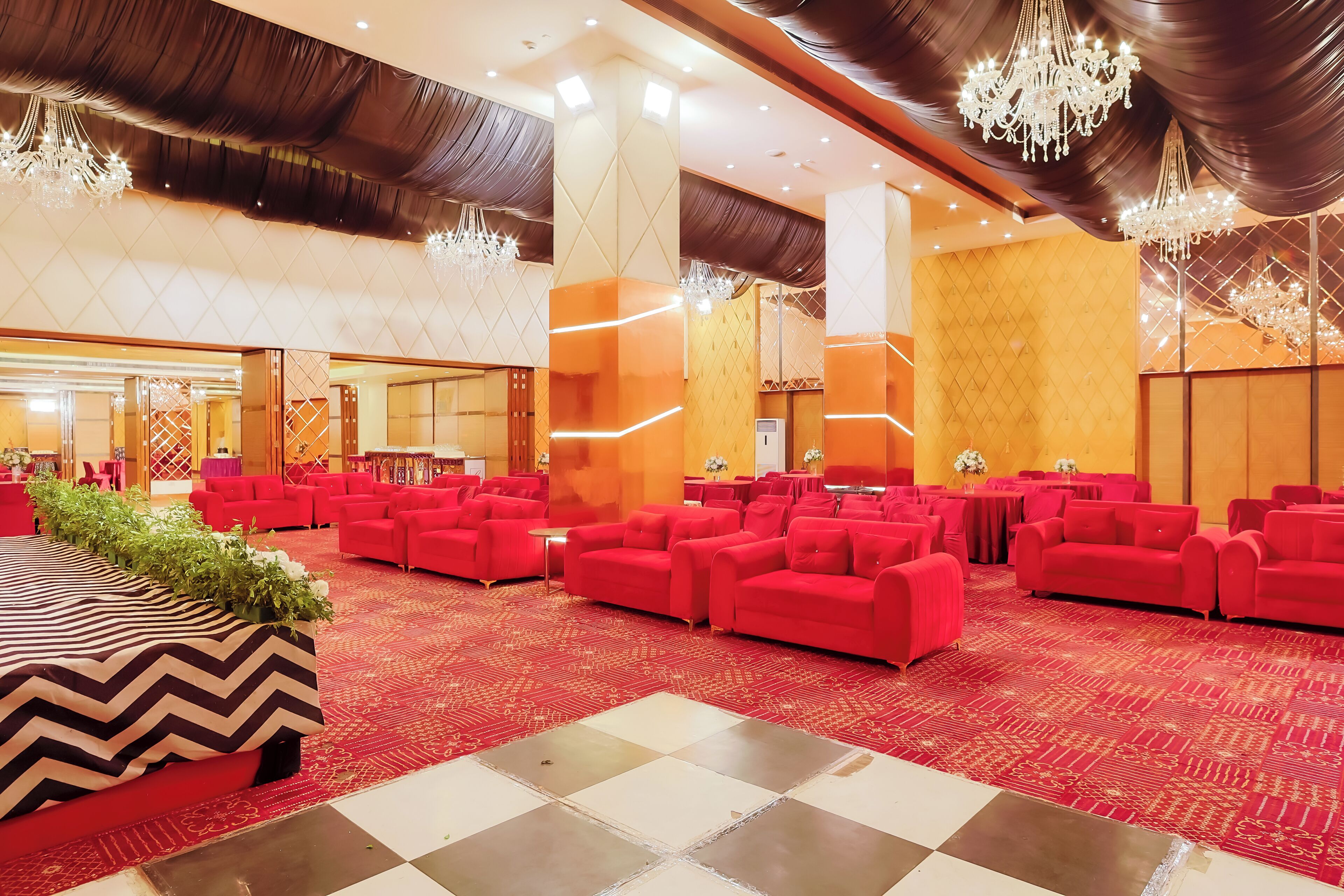A spacious hall with red sofas arranged in rows, a patterned carpet, and a chandelier.