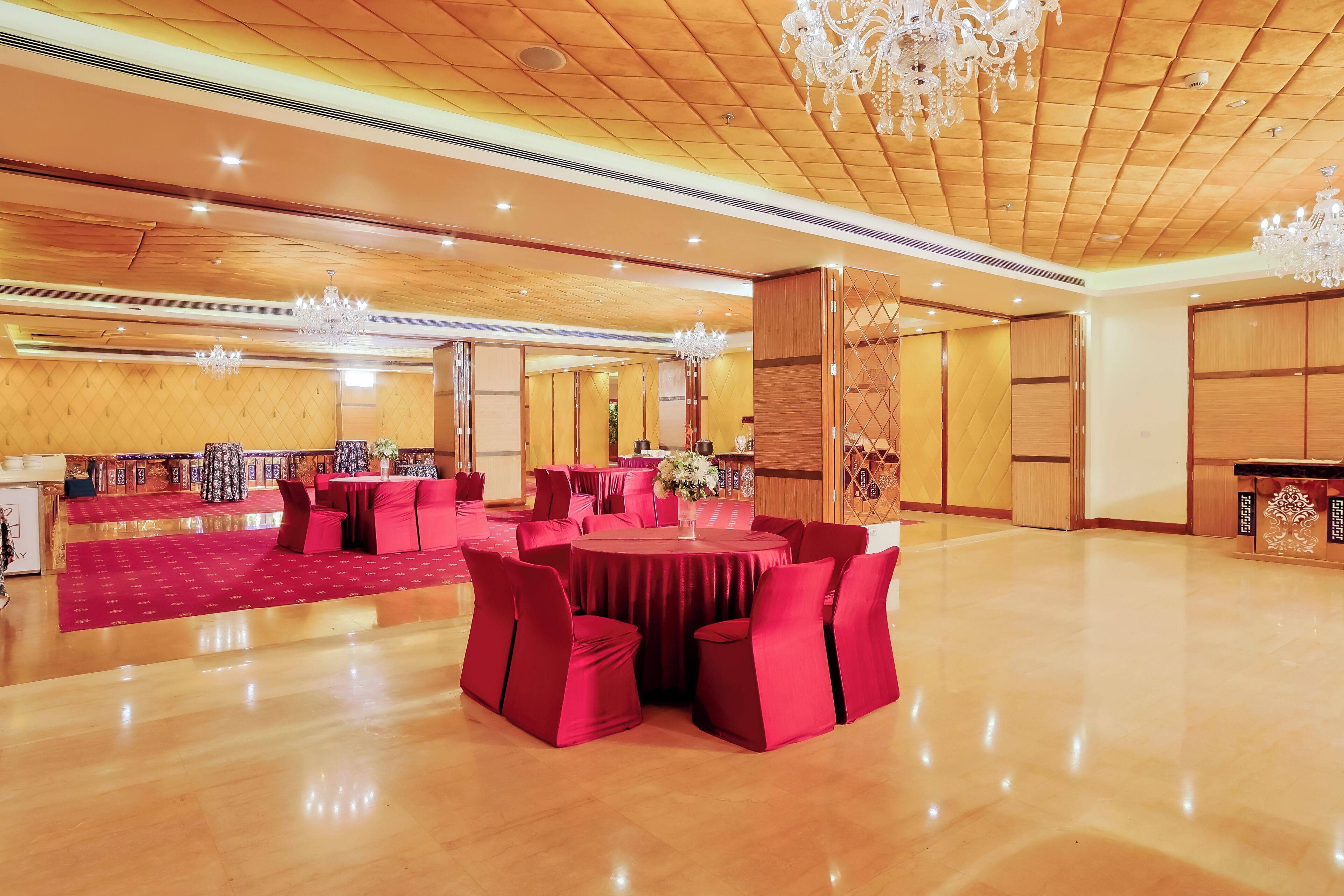 A spacious banquet hall with round tables and red chairs, wooden paneling, and chandeliers.