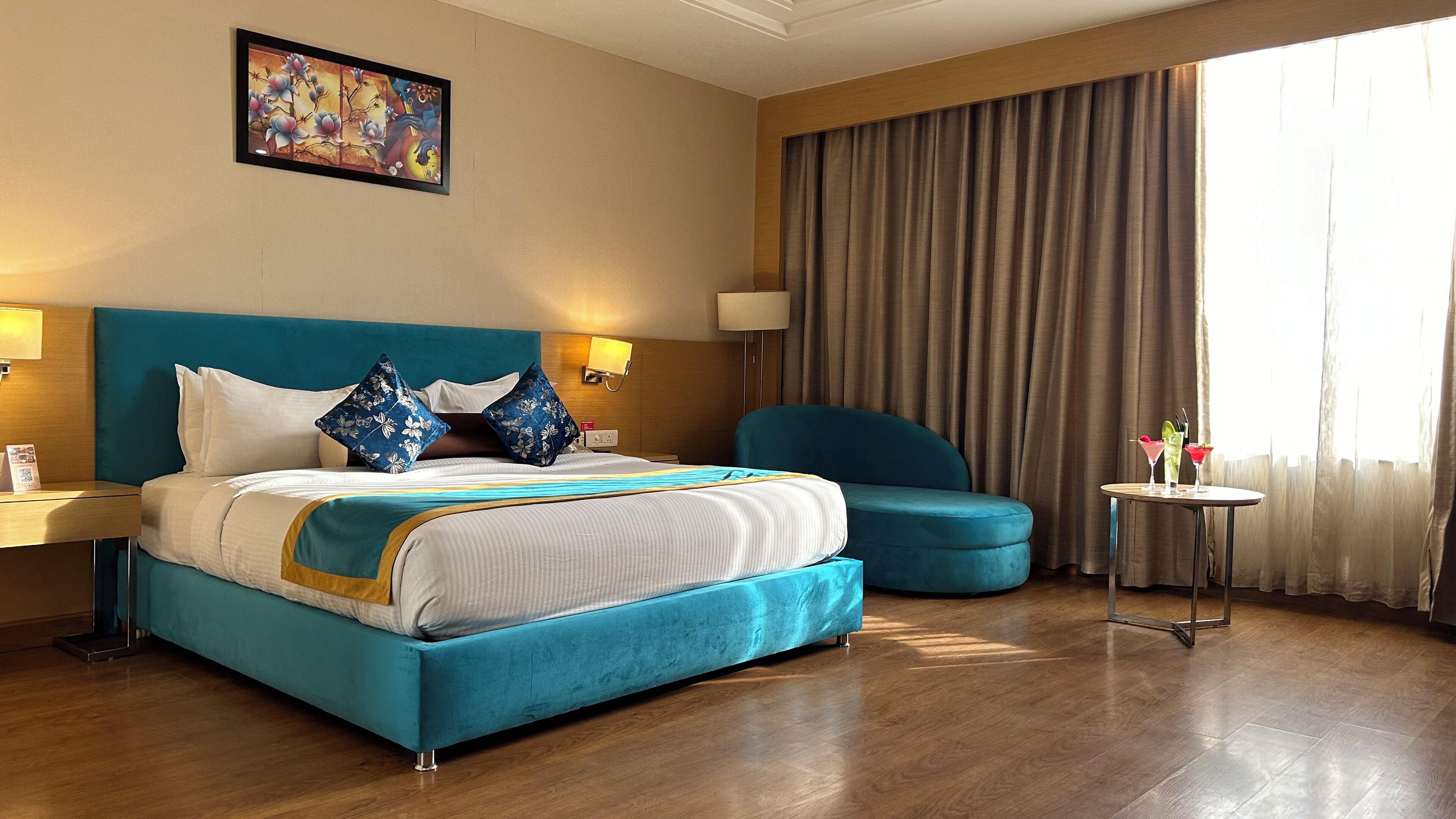 A hotel room with a large bed, a blue armchair, a small table with drinks, and a painting on the wall.