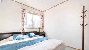 A neatly made bed with a blue blanket and pillows, a wooden coat rack, and a window with floral curtains. - 1 bedroom apartment in saitama (川口市西川口)
