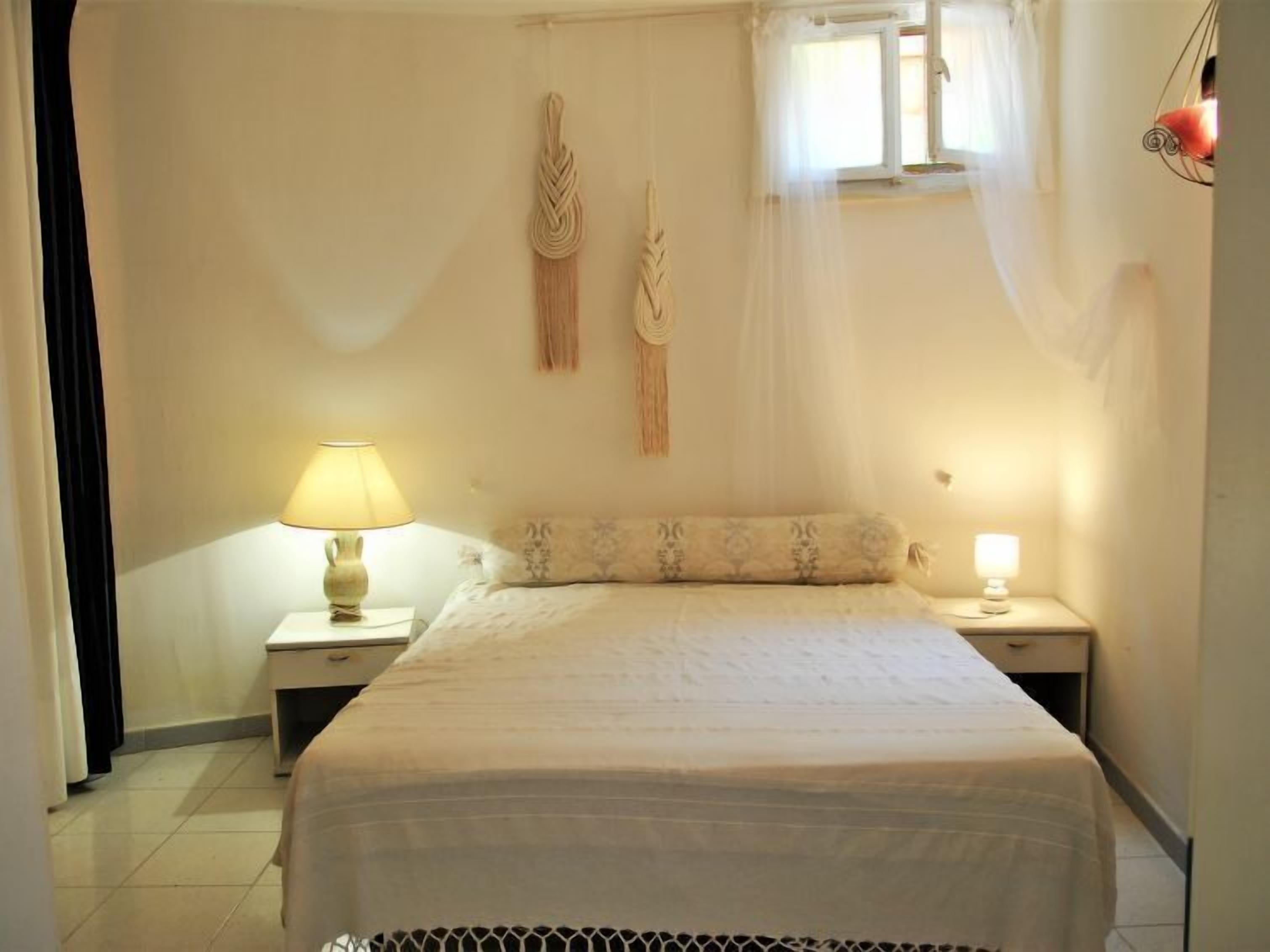 Une chambre à coucher avec un lit, deux tables de chevet, une lampe sur chaque table, une fenêtre avec des rideaux légers et une décoration murale en macramé.