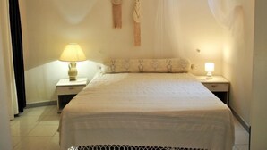 A bedroom with a bed, two bedside tables, a lamp on each table, a window with sheer curtains, and a wall-mounted macrame piece.
