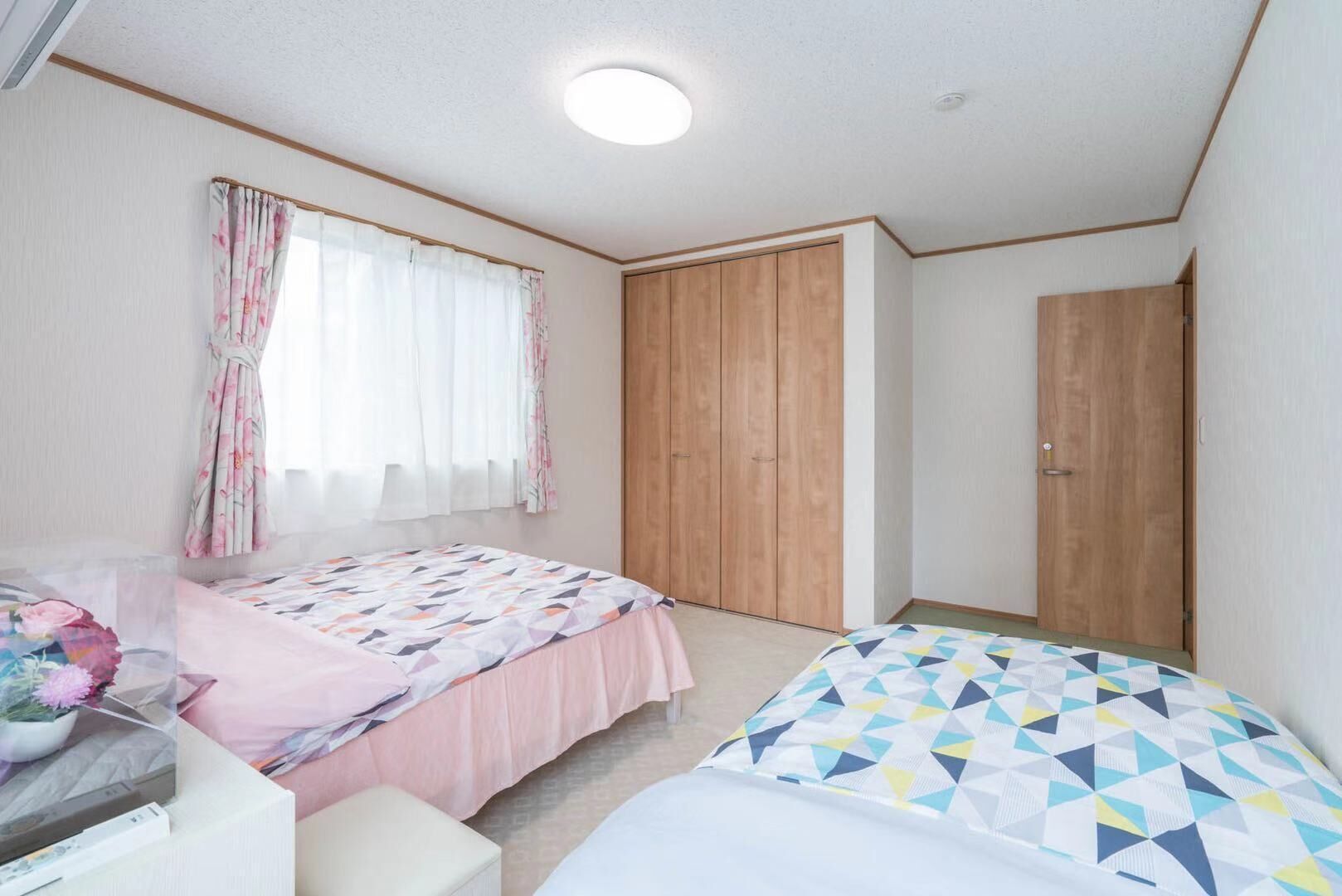 A bedroom with two beds, a wooden wardrobe, a window with curtains, and a ceiling light.