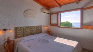 Une chambre avec un plafond en bois, un lit recouvert d’une couvre-lit à motifs floraux, un climatiseur fixé au mur et une fenêtre donnant sur un paysage verdoyant et la mer.