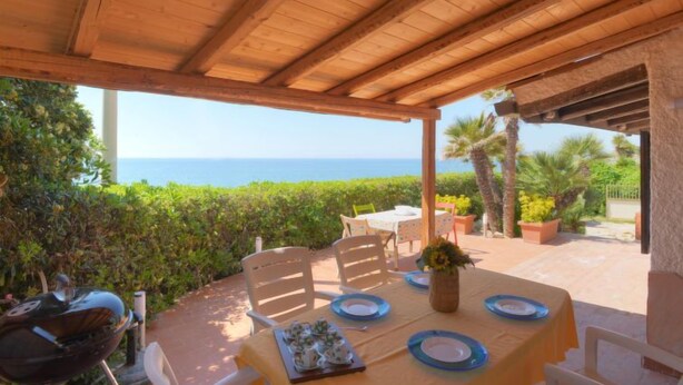 Un espace extérieur abrité, aménagé pour les repas, avec vue sur la mer, comprenant des tables et des chaises en bois, ainsi qu’un barbecue.