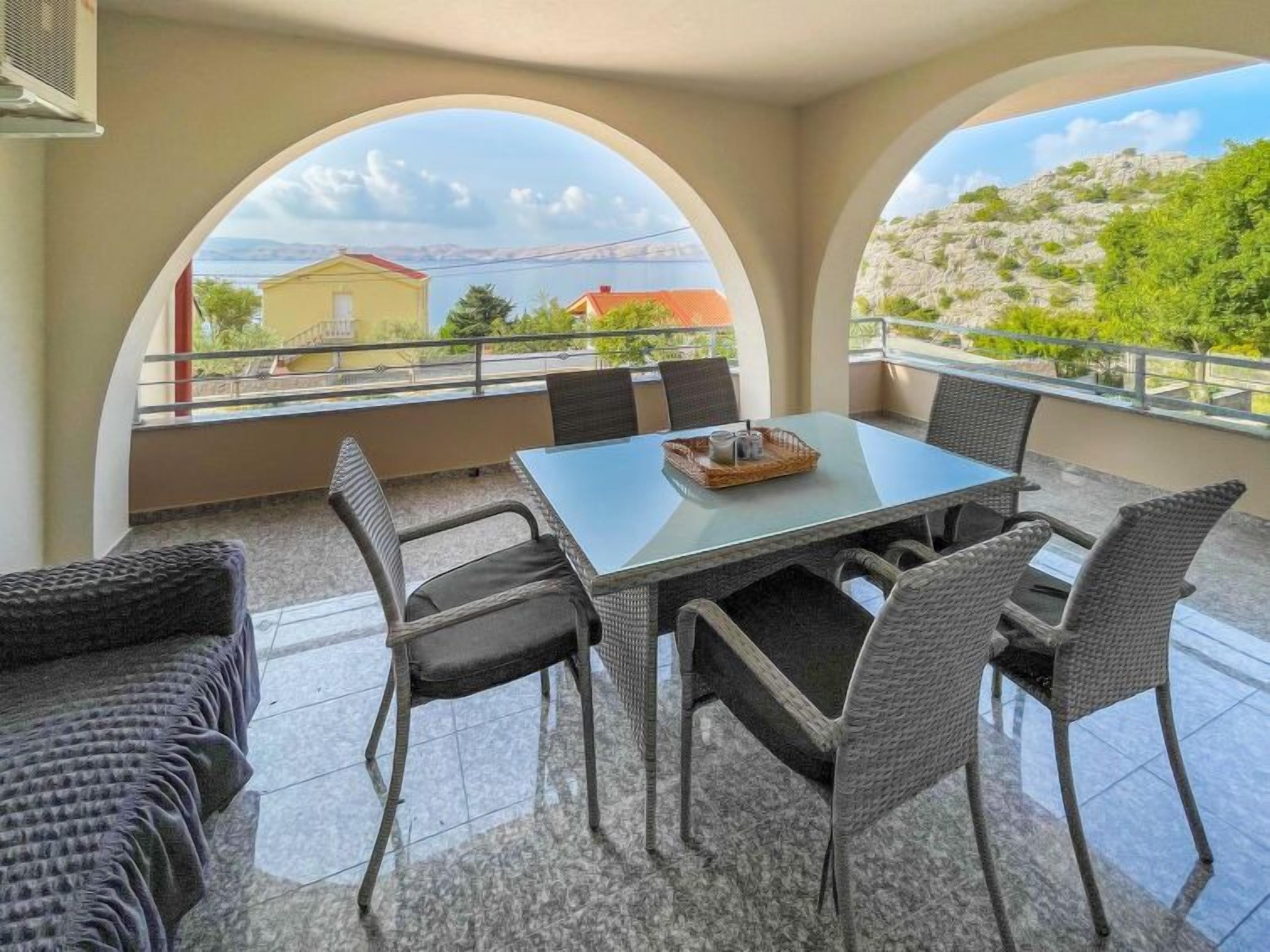 Ein Balkon mit Glastisch und Korbstühlen, der einen schönen Ausblick auf eine Landschaft mit Gebäuden und Grünflächen bietet.
