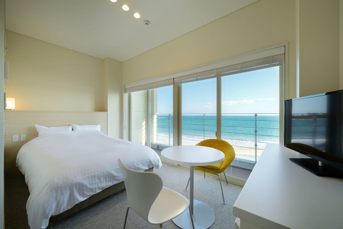 A hotel room with a bed, a round table, a chair, a TV, and a large window with a view of the beach.