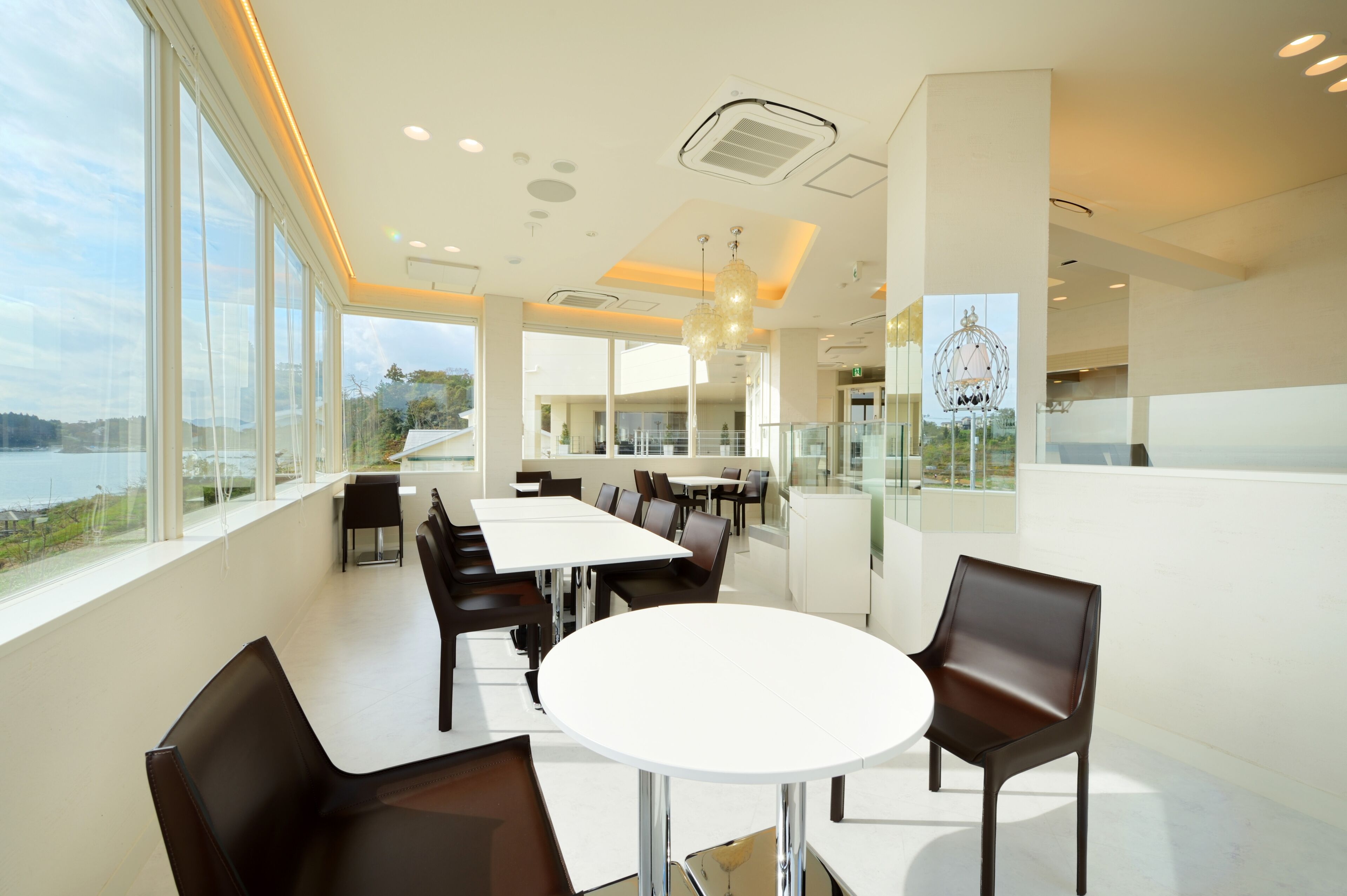 Un espace repas moderne avec des tables blanches et des chaises marron, de grandes fenêtres offrant une vue sur la mer et un lustre décoratif.