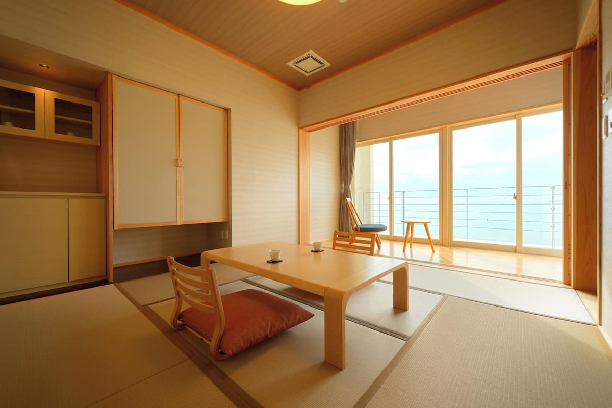 A traditional Japanese room with tatami flooring, a low wooden table, and sliding doors leading to a balcony with a view of the sea.