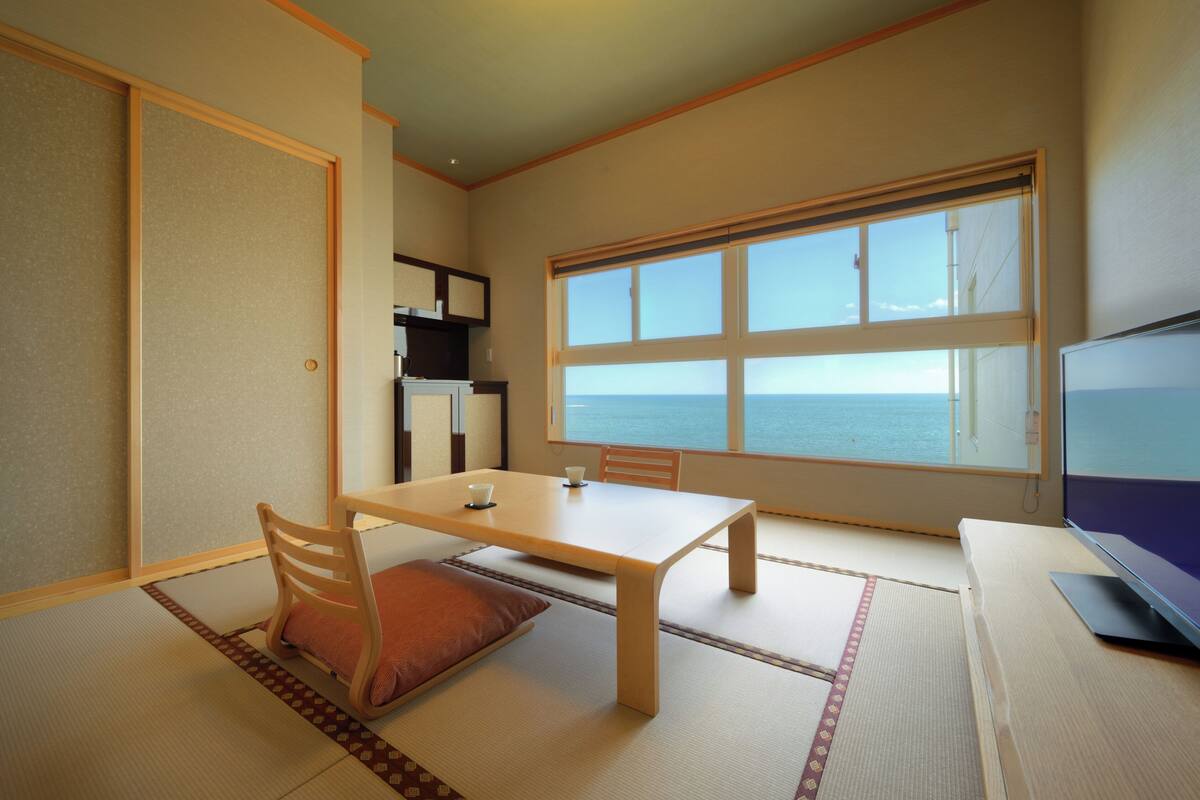 A room with tatami flooring, a low table with chairs, a television, and a large window with a view of the sea.