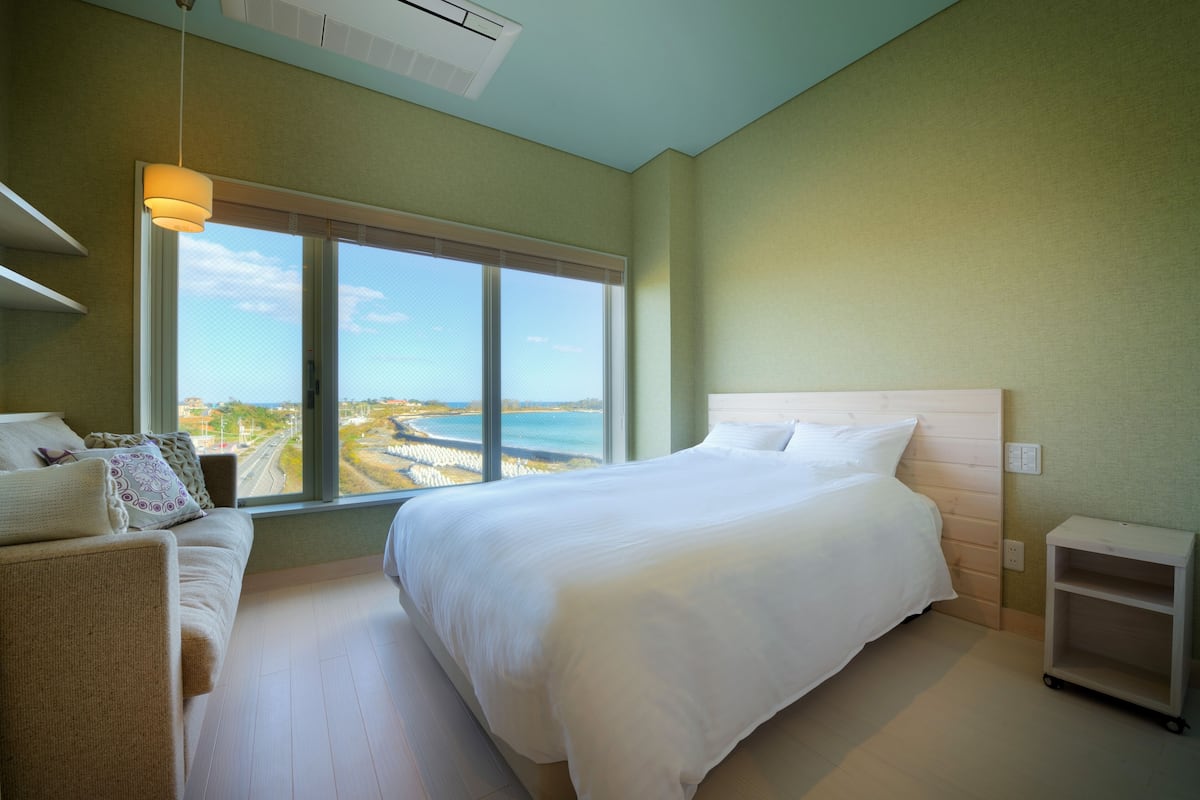 A bedroom with a large bed, a sofa, a view of the sea, and a ceiling fan.