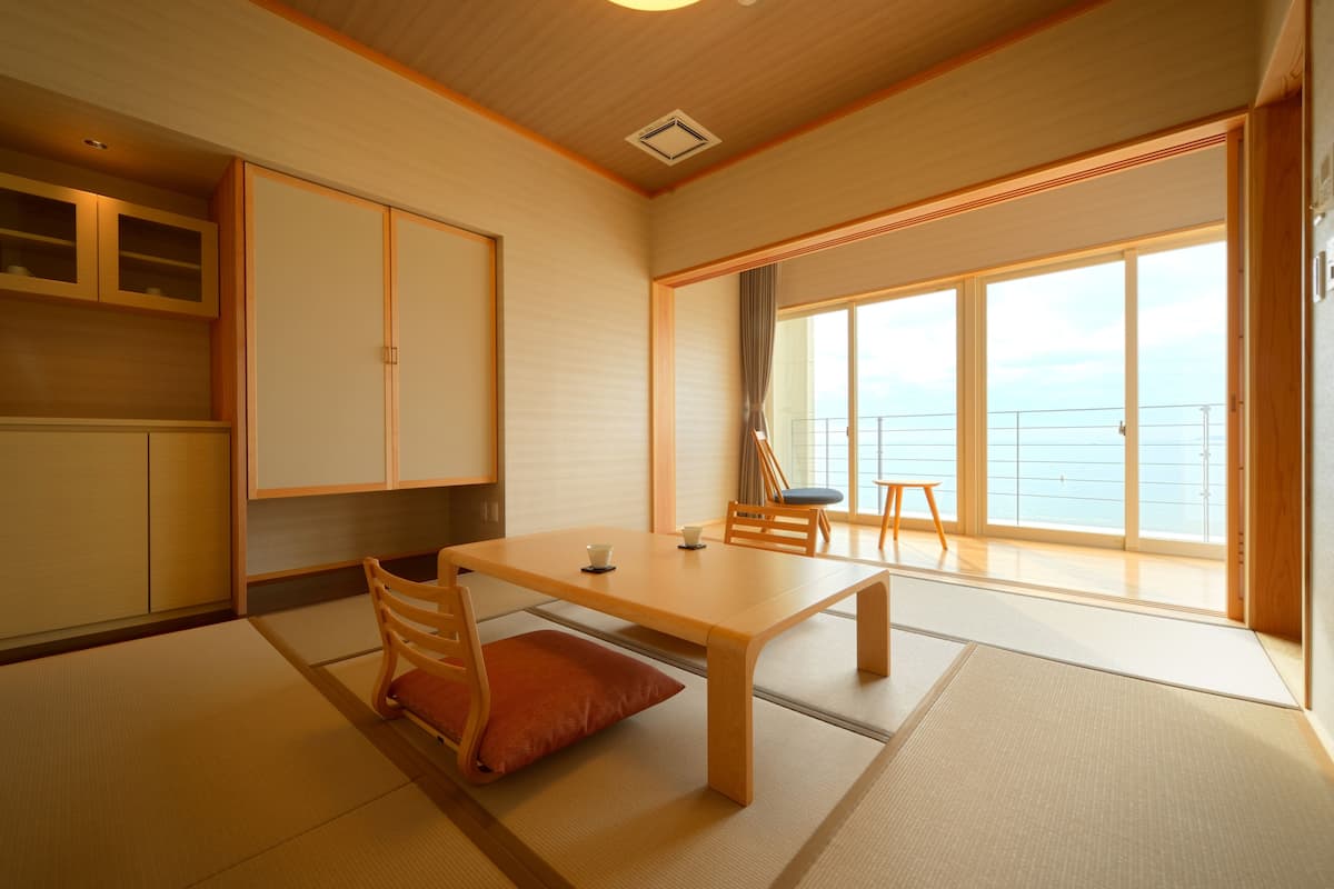 A traditional Japanese room with tatami flooring, a low wooden table, and sliding doors leading to a balcony with a view of the sea.