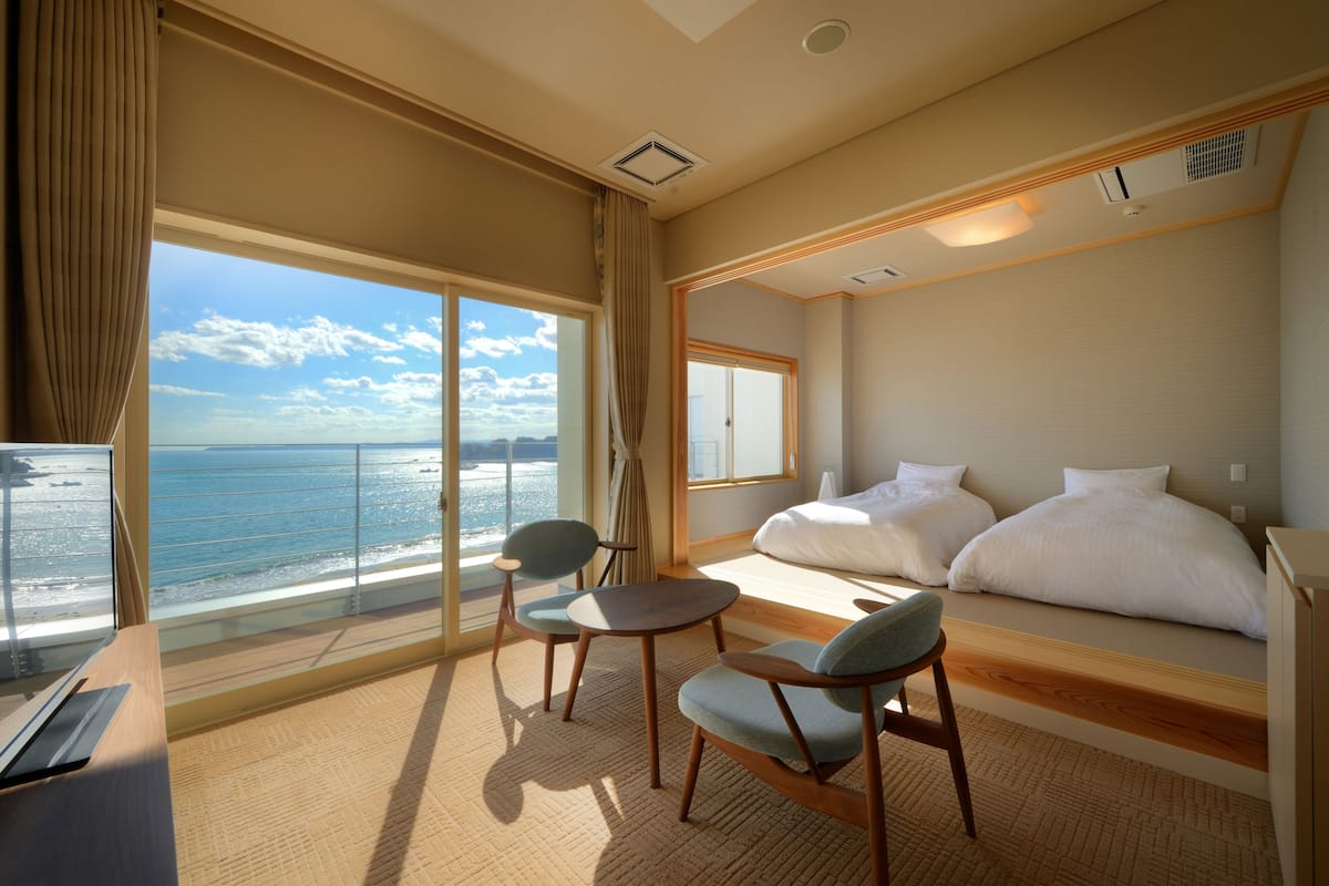 A hotel room with a large window overlooking the sea, a bed, a desk, and two chairs.