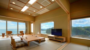 Een kamer met tatami matten, een houten tafel, stoelen, een televisie en grote ramen met uitzicht op zee.