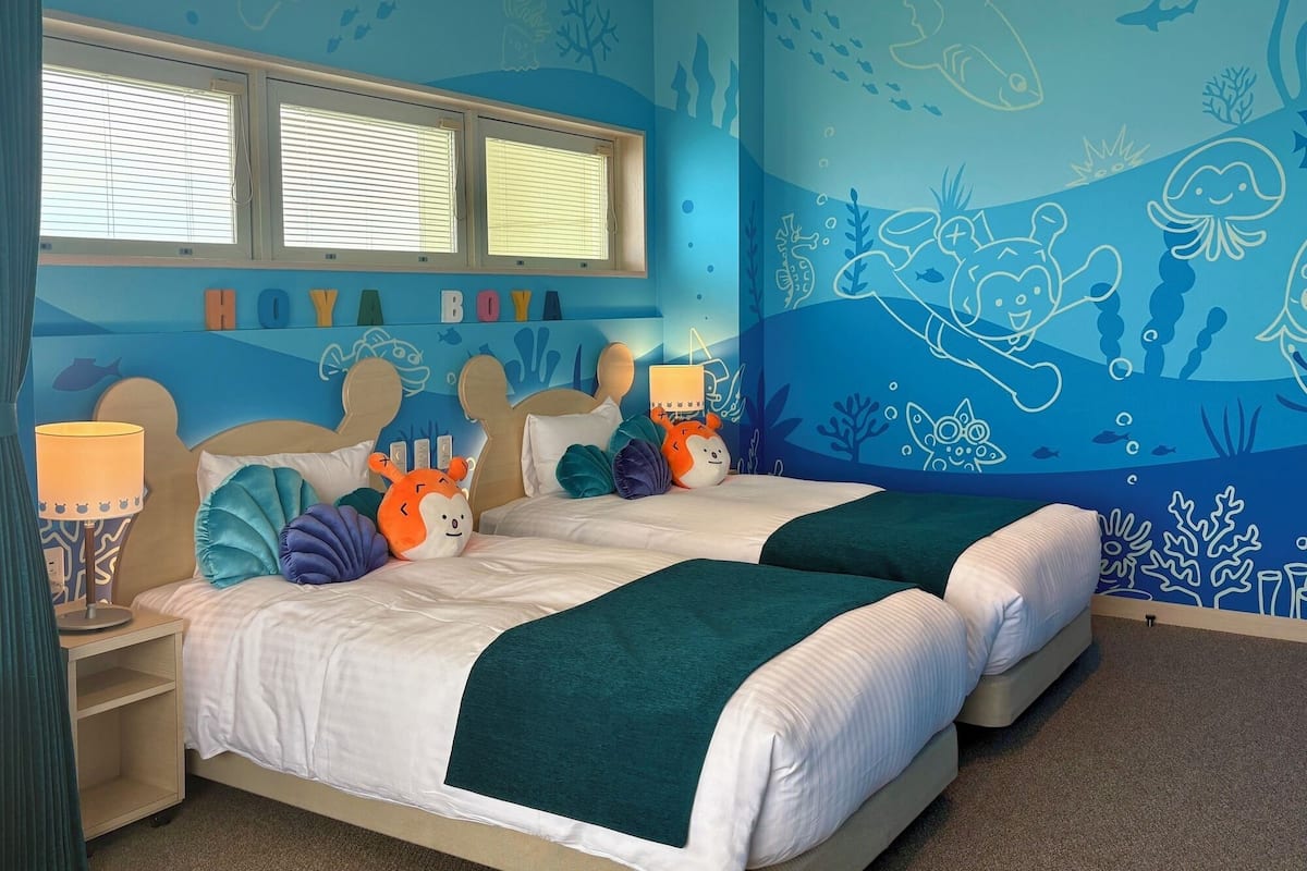 A room with two beds, a window with blinds, and a wall mural with underwater themes.