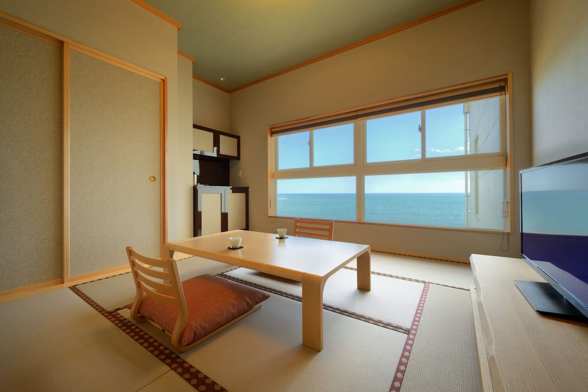 A room with tatami flooring, a low table with chairs, a television, and a large window with a view of the sea.