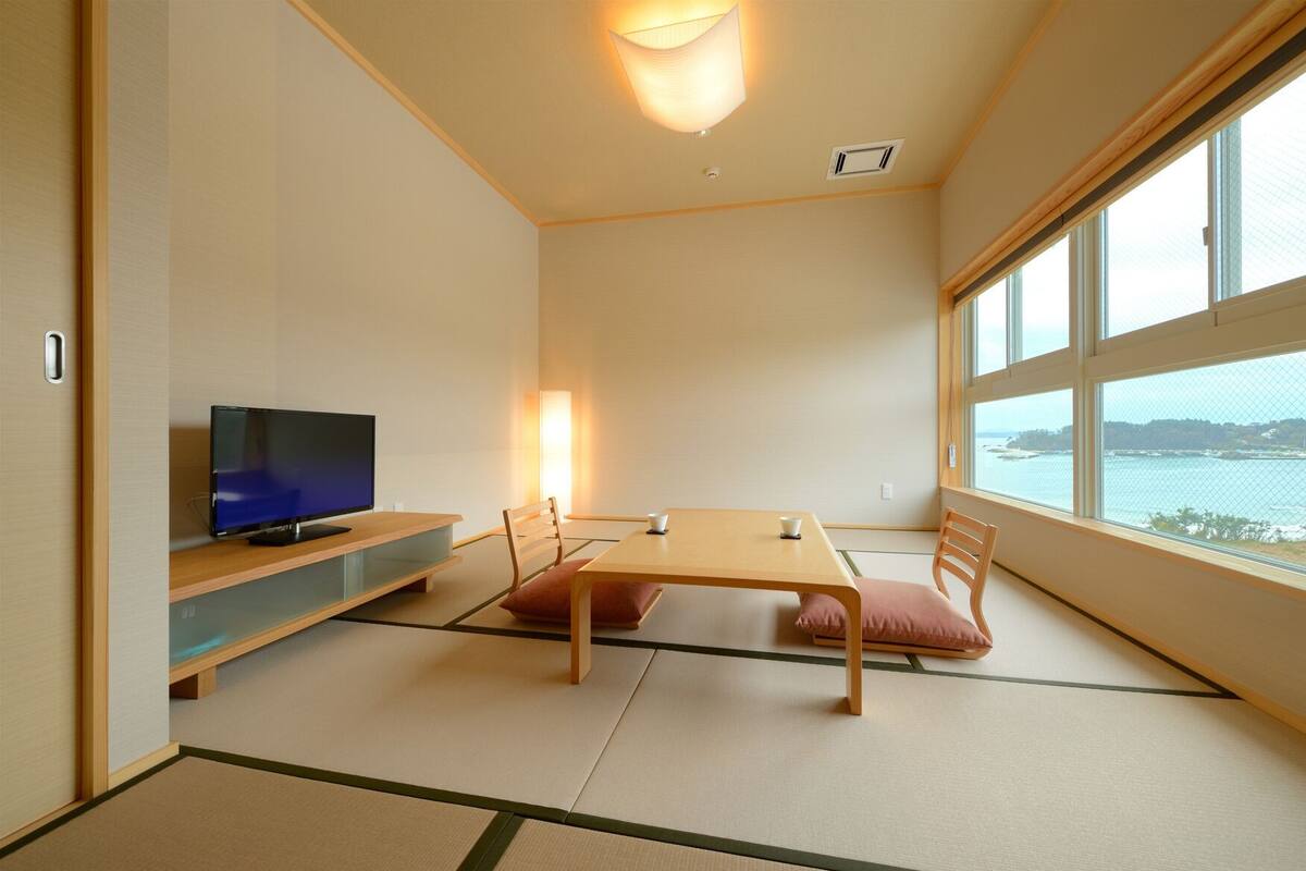 A room with tatami flooring, a television on a low shelf, a table with chairs, and a large window with a view of the sea.