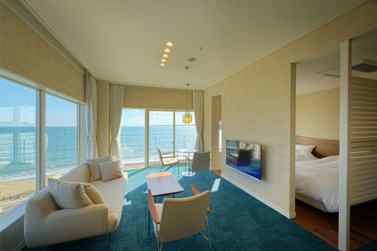 A hotel room with a large window offering a sea view, a sofa, a dining table, and a bed.