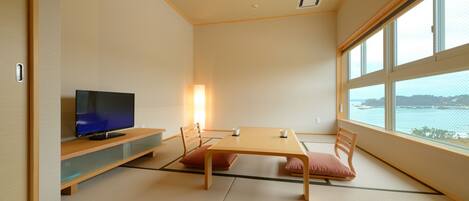 A traditional Japanese room with tatami flooring, a television on a low shelf, a table with cushions, and a large window offering a view of the sea.