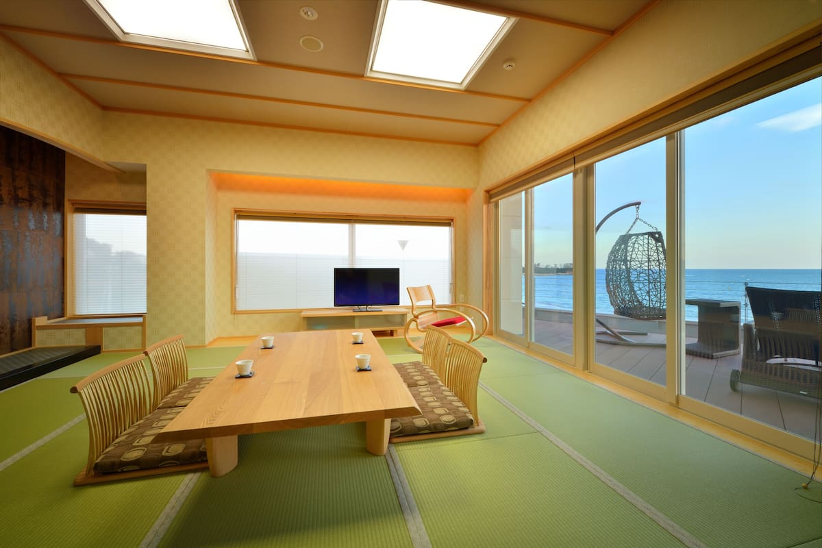A room with tatami mats, a wooden table, tatami seating, a television, and large windows overlooking the sea.