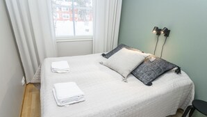 Una cama bien hecha con un edredón blanco acolchado, dos almohadas y toallas blancas dobladas. Una silla negra está ubicada al pie de la cama.