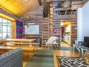 A log cabin interior with a wooden table, chairs, a ladder, and a kitchen area.