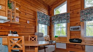 A cozy cabin interior with wooden walls, a dining area with a round table and chairs, a kitchen area with a coffee maker, and large windows with patterned curtains. - Pikku-veke by Interhome (Inari)