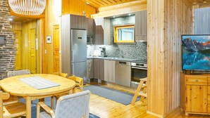 Une cuisine chaleureuse et bien éclairée, avec des meubles en bois, un réfrigérateur et un four à micro-ondes. On y trouve un coin repas avec une table et des chaises en bois, ainsi qu’un téléviseur fixé au mur.